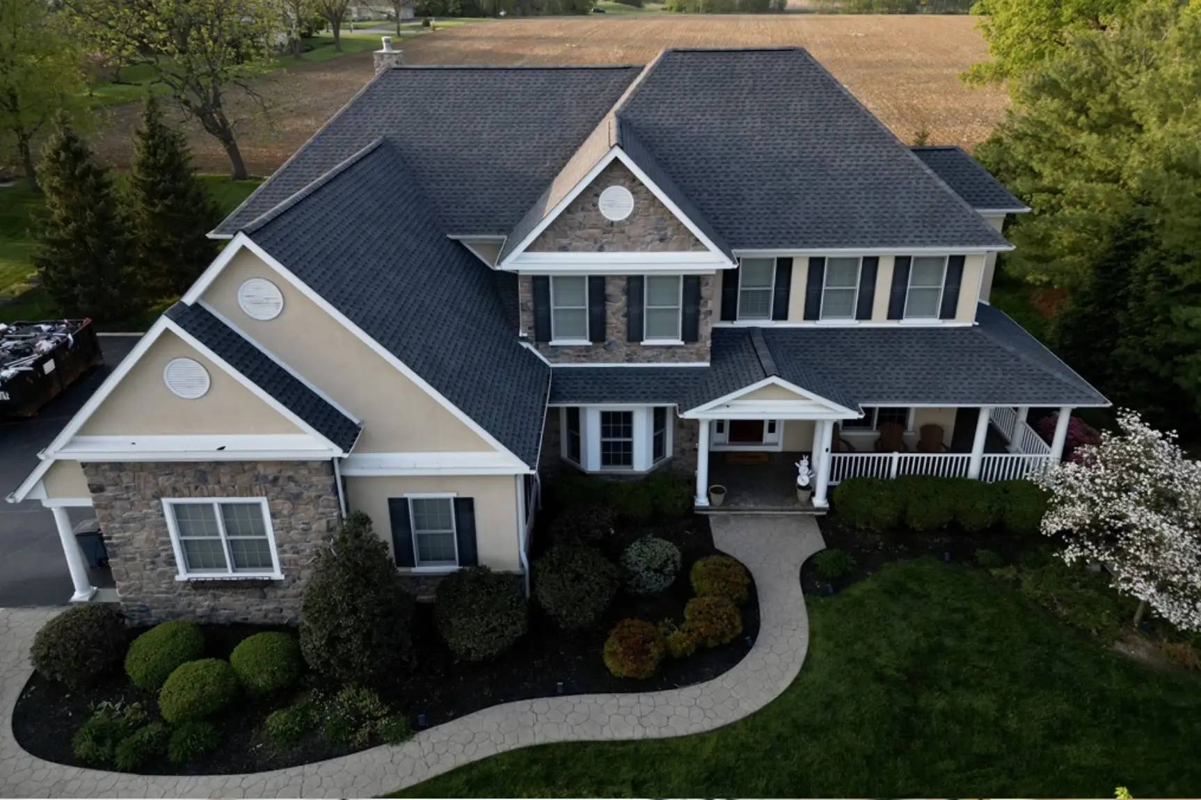 Drone shot of beige house with dark gray asphalt shingle roof. Large two-story house with vinyl and stone facade. Landscaped front yard and large field in back with tall trees on either side.