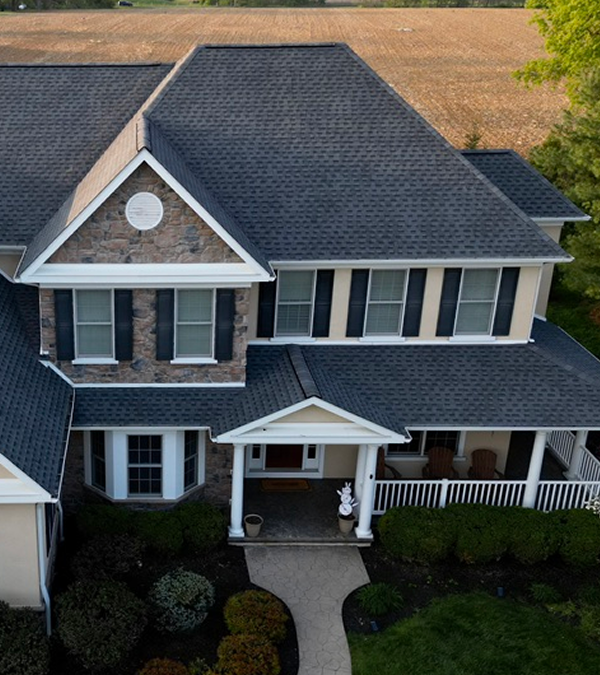 A large suburban house with a grey roof and stone facade, surrounded by manicured gardens and pathway. Open fields and trees lie in the distant background.